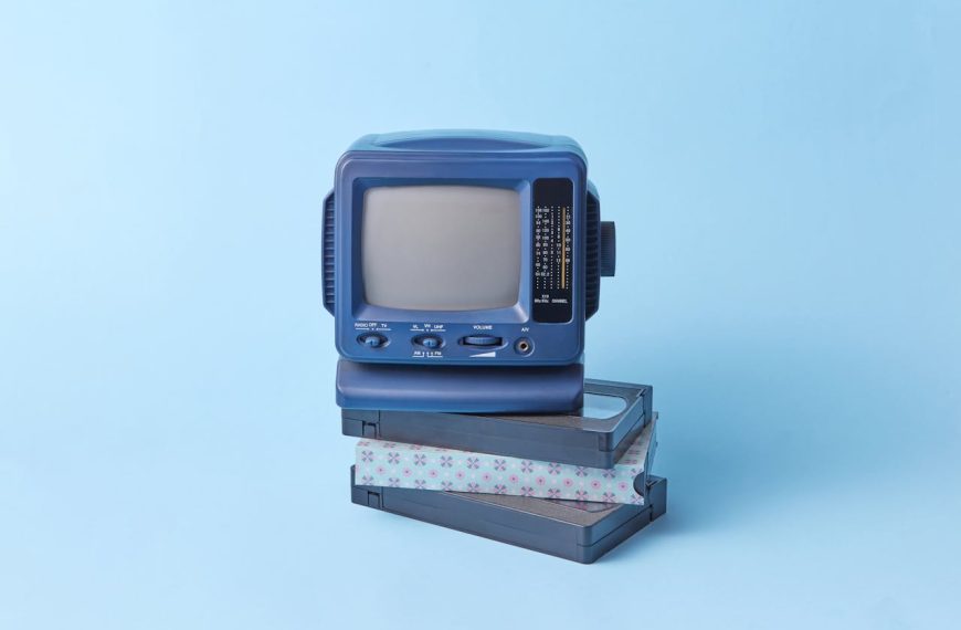 Small vintage blue television with dials, placed on stacked VHS tapes, set against light blue background