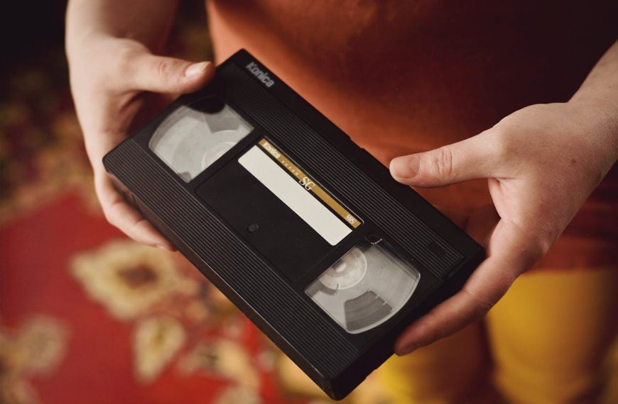 Close-up of hands holding a VHS videocassette tape. The black plastic case displays a white label area in the center, positioned above a patterned carpet or rug with reddish tones