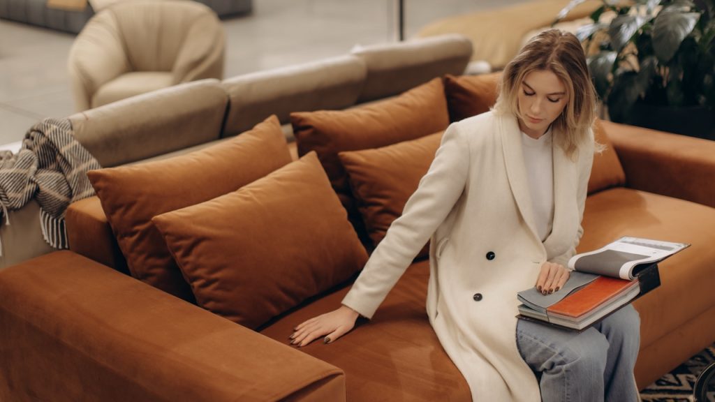 A woman in a white coat inspects a contemporary orange velvet sofa at a furniture showroom.