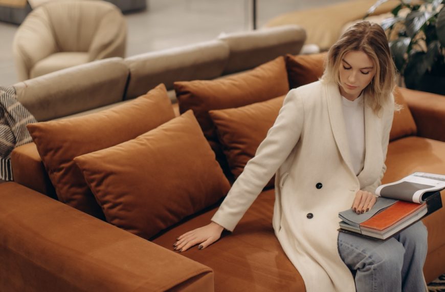 A woman in a white coat inspects a contemporary orange velvet sofa at a furniture showroom.