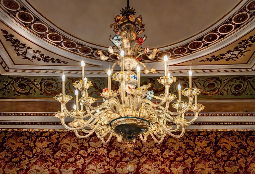 Ornate crystal chandelier, multiple candle-style bulbs, gold detailing, suspended from a decorated ceiling, surrounded by intricate crown molding, red and gold patterned wallpaper background