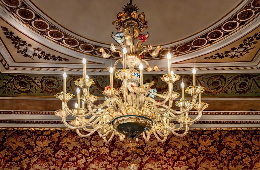 Ornate crystal chandelier, multiple candle-style bulbs, gold detailing, suspended from a decorated ceiling, surrounded by intricate crown molding, red and gold patterned wallpaper background