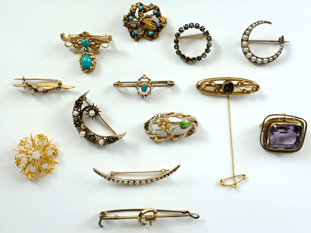Collection of antique brooches and pins, various shapes and styles, featuring gold, gemstones, pearls, and intricate metalwork, arranged on a white background
