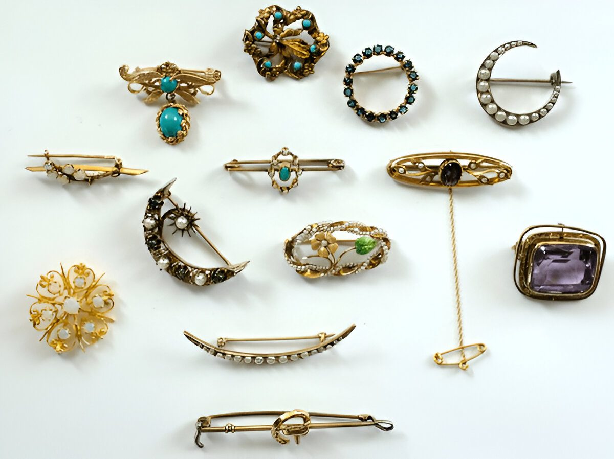 Collection of antique brooches and pins, various shapes and styles, featuring gold, gemstones, pearls, and intricate metalwork, arranged on a white background