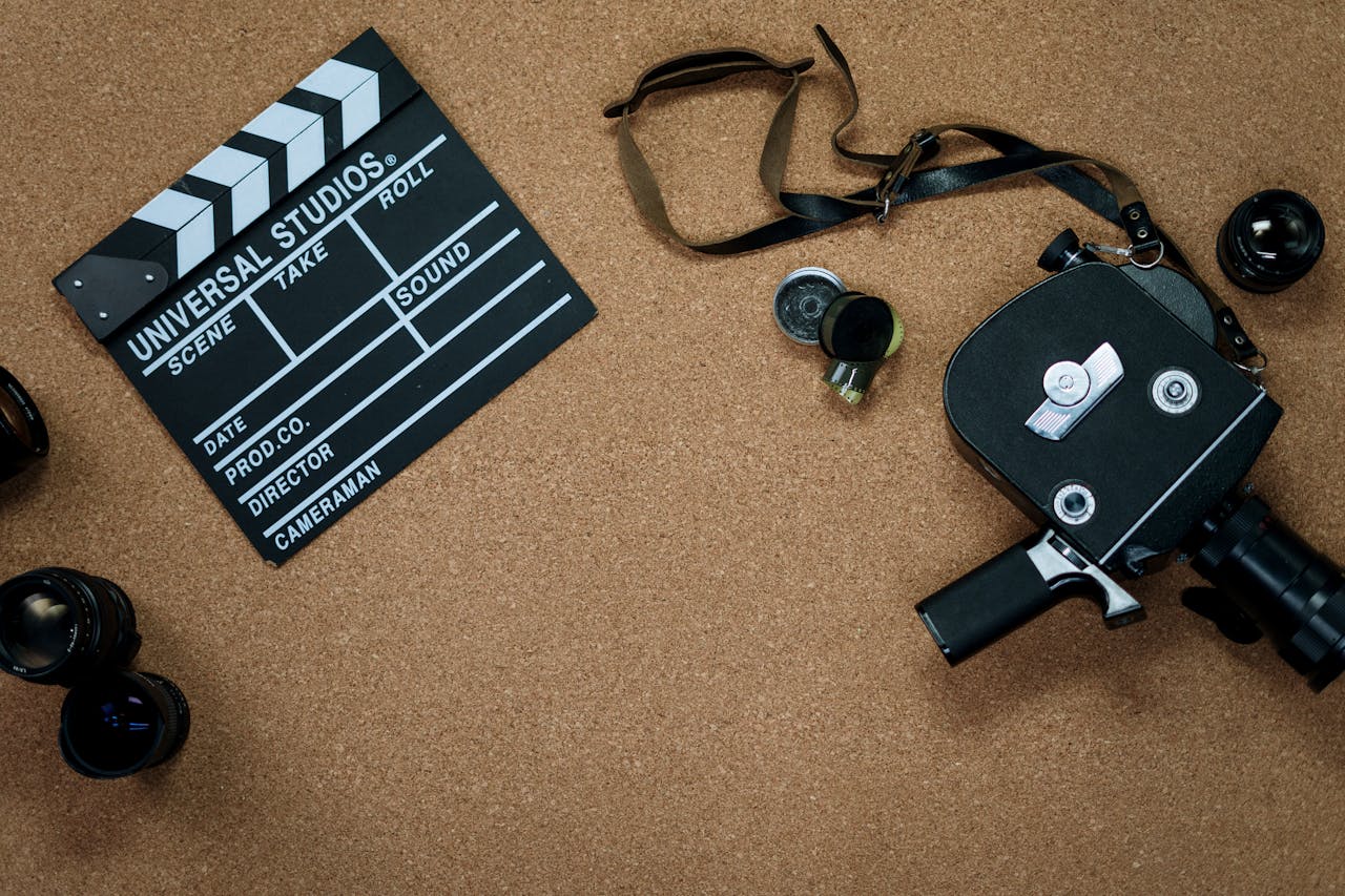 Film production equipment on cork surface featuring a Universal Studios clapperboard, vintage 8mm or 16mm camera with strap, camera lens, and viewfinder. Classic filmmaking tools arranged for nostalgic composition