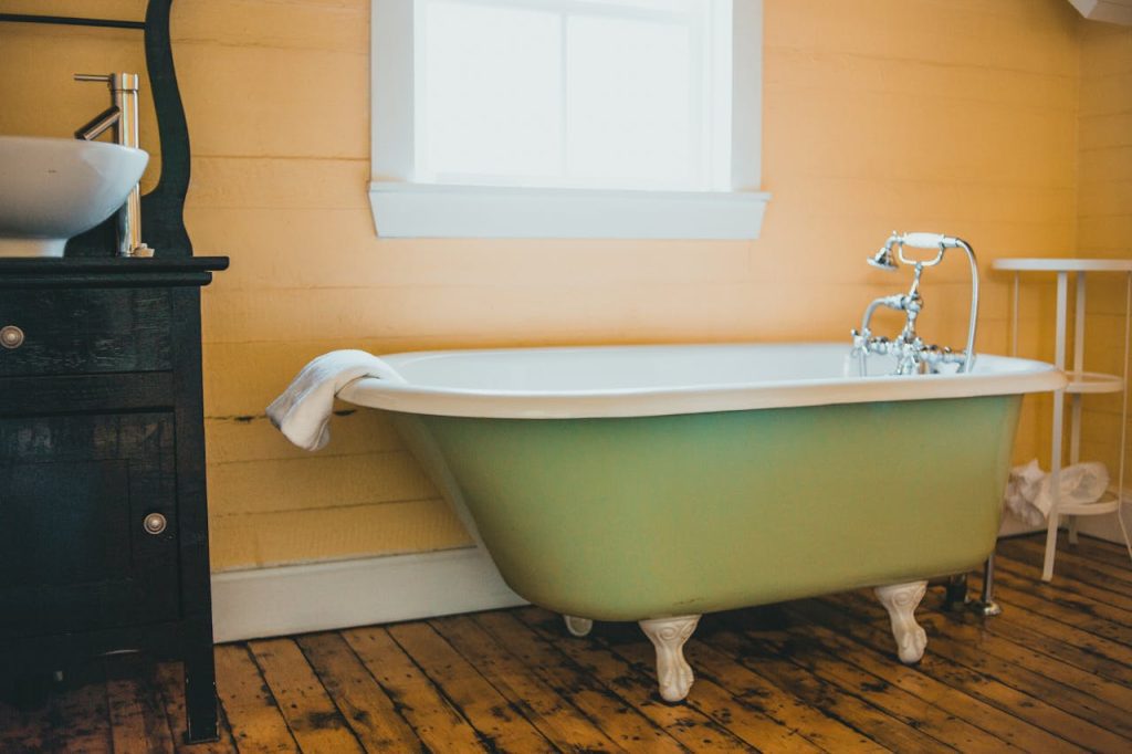 Vintage-style bathroom, green clawfoot bathtub, white towel draped on tub, wooden floor, yellow paneled wall, window above tub, black cabinet with basin sink, white metal stand beside tub