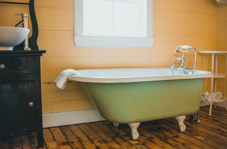 Vintage-style bathroom, green clawfoot bathtub, white towel draped on tub, wooden floor, yellow paneled wall, window above tub, black cabinet with basin sink, white metal stand beside tub