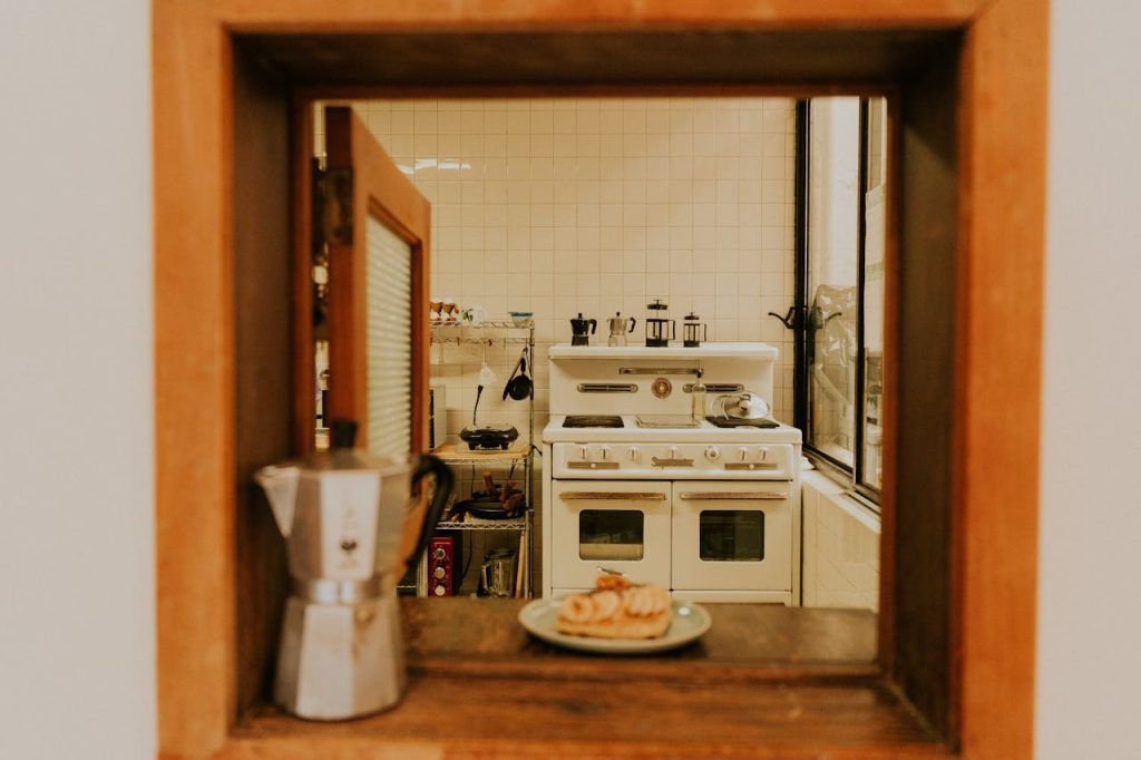A vintage kitchen is viewed through a wooden-framed pass-through window, featuring a classic white stove, tiled walls, and French press coffee makers. A stovetop espresso maker and a plate of pastries rest on the wooden ledge