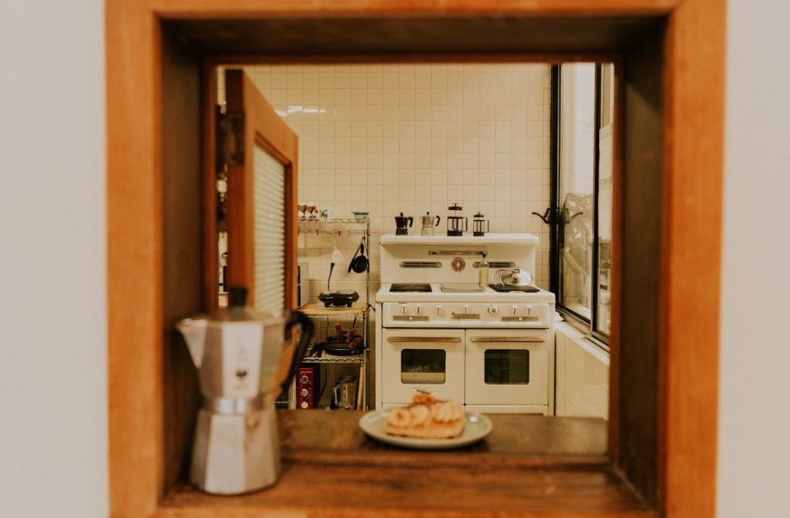 A vintage kitchen is viewed through a wooden-framed pass-through window, featuring a classic white stove, tiled walls, and French press coffee makers. A stovetop espresso maker and a plate of pastries rest on the wooden ledge