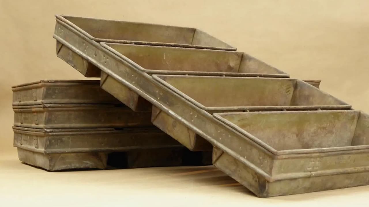 Weathered metal baking pans stacked against a beige background. Several rectangular loaf pans are arranged diagonally, while others are stacked horizontally. The industrial-style pans show signs of age and use
