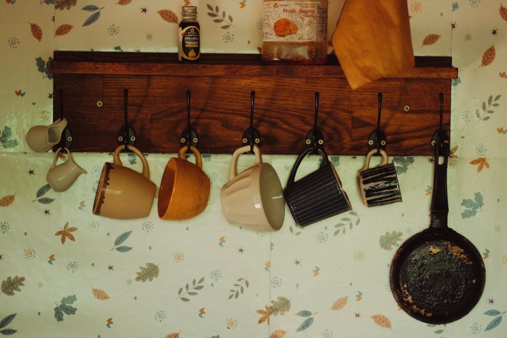 Wooden coat rack with coffee mugs hanging from hooks against leaf-patterned wallpaper. Various mugs in beige, orange, and striped designs. A cast iron pan hangs on the right, with small bottles on the shelf above