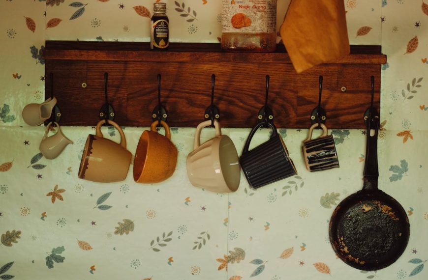 Wooden coat rack with coffee mugs hanging from hooks against leaf-patterned wallpaper. Various mugs in beige, orange, and striped designs. A cast iron pan hangs on the right, with small bottles on the shelf above