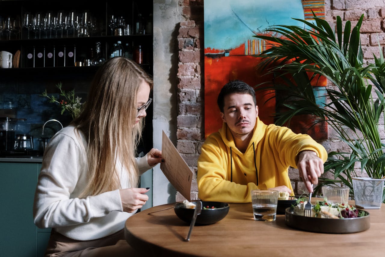 Man and woman sitting at a wooden table, man wearing a yellow hoodie reaching for food with a fork, bowls of food and glasses of water on the table