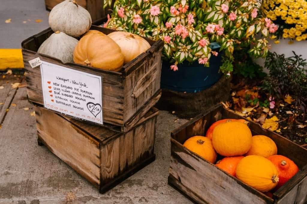 Wooden crates of autumn squash varieties at farmers market, sign listing New England pie pumpkin and butternut squash, orange pumpkins in separate crate, pink and yellow flowers nearby