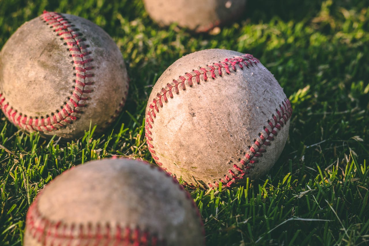 Worn baseballs, red stitching, placed on green grass, signs of dirt and heavy use, close-up outdoor setting