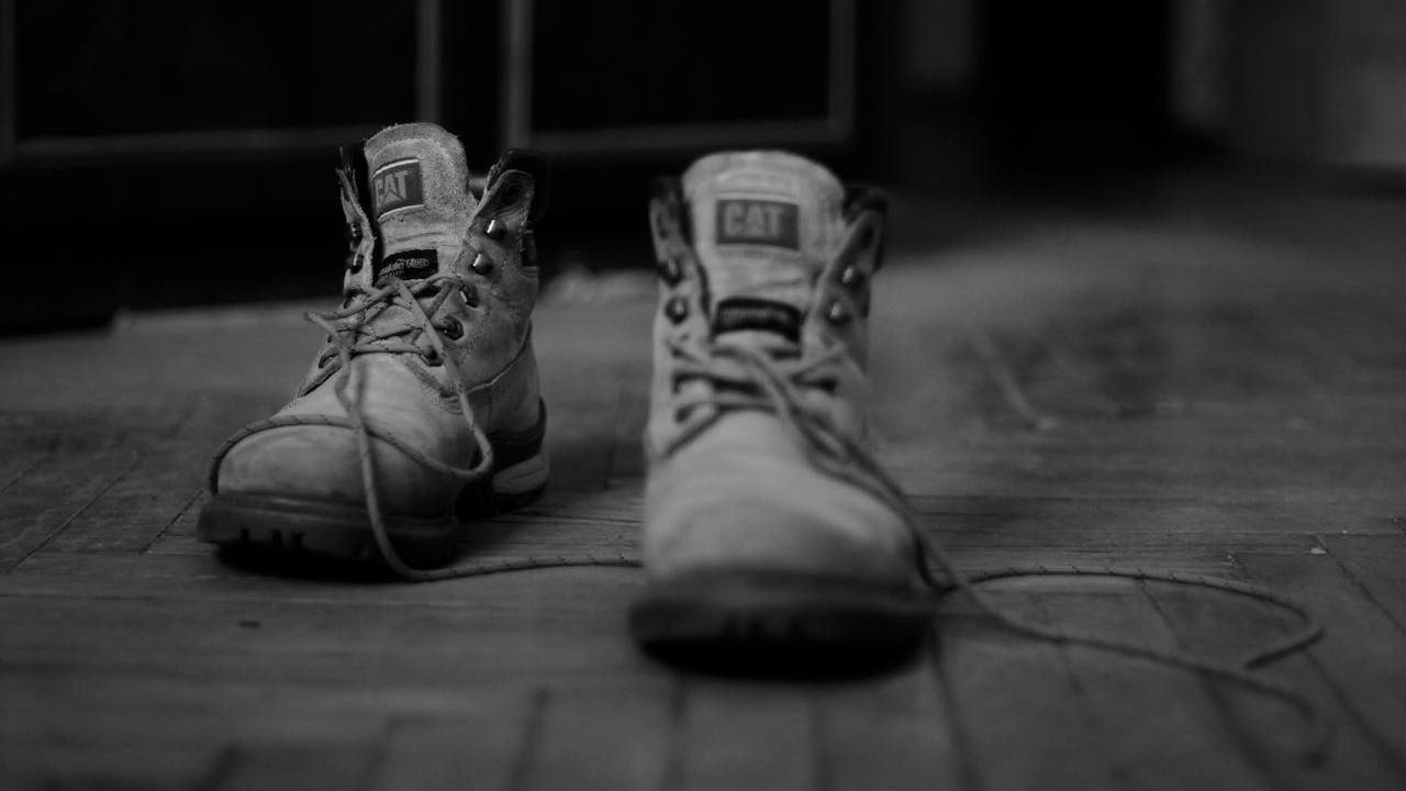 Pair of worn-out CAT work boots, untied laces, resting on a wooden floor, captured in black and white
