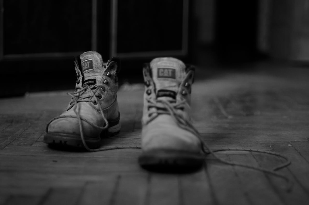 Pair of worn-out CAT work boots, untied laces, resting on a wooden floor, captured in black and white