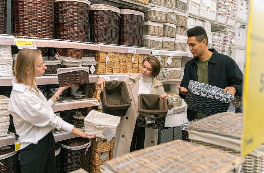 Three people shopping for storage baskets in a home goods store. They're examining different wicker baskets in various colors and styles. Store shelves are stocked with numerous basket options in brown, white, and patterned designs