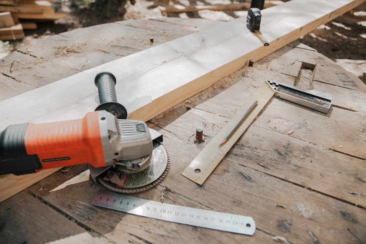 Angle grinder with an orange handle, metal rulers, wooden plank, tape measure, pencil outdoor woodworking setup
