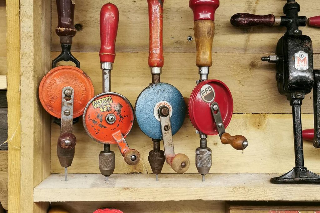 Close-up of vintage hand drills, various colors and brands, wooden and metal handles, mounted on a wooden wall
