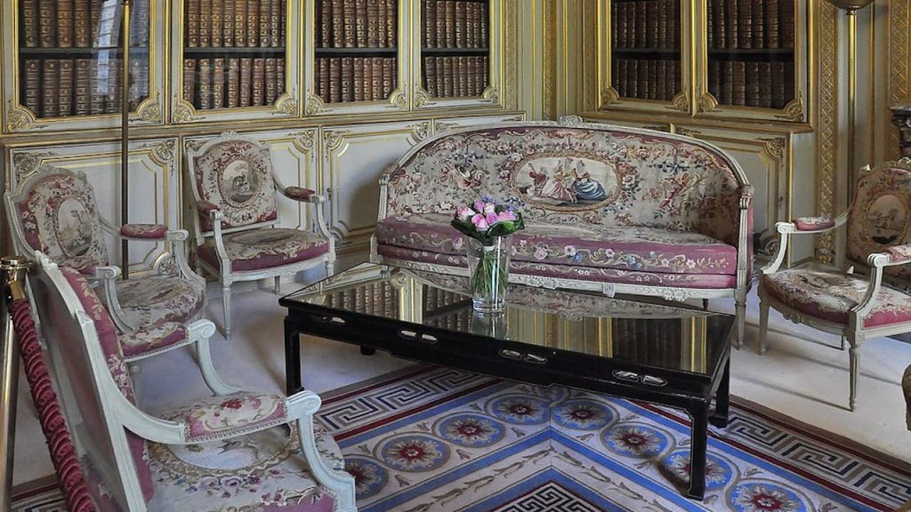 Opulent French rococo salon featuring cream and gold paneled walls with built-in bookshelves. White-framed settee and armchairs with pink tapestry upholstery surround a black lacquered coffee table on an ornate geometric-patterned rug