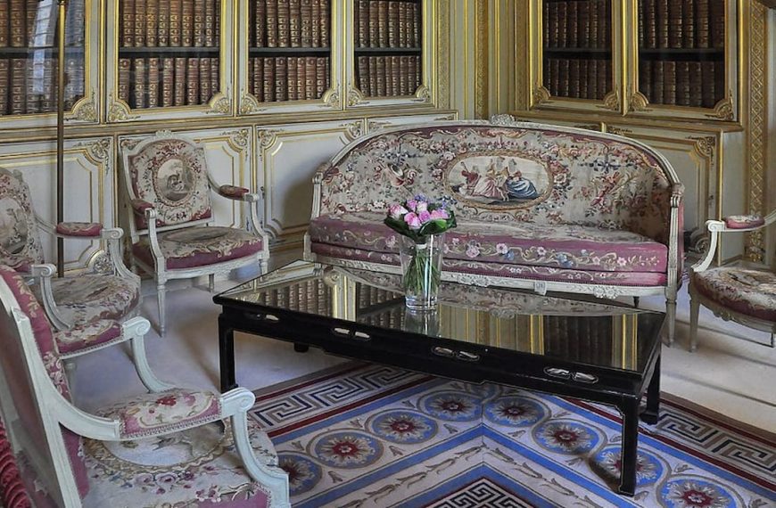 Opulent French rococo salon featuring cream and gold paneled walls with built-in bookshelves. White-framed settee and armchairs with pink tapestry upholstery surround a black lacquered coffee table on an ornate geometric-patterned rug