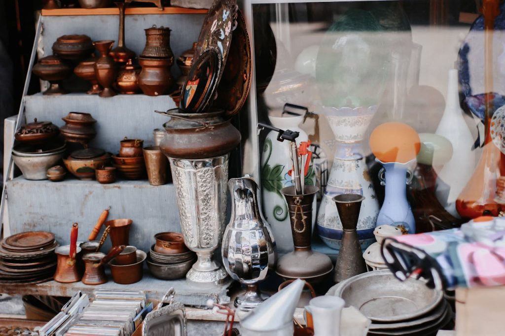 assortment of antique metalware, ceramics, and glass items displayed on shelves and tables at a vintage market