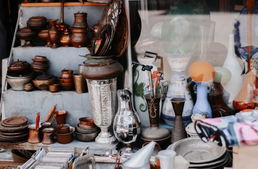 assortment of antique metalware, ceramics, and glass items displayed on shelves and tables at a vintage market