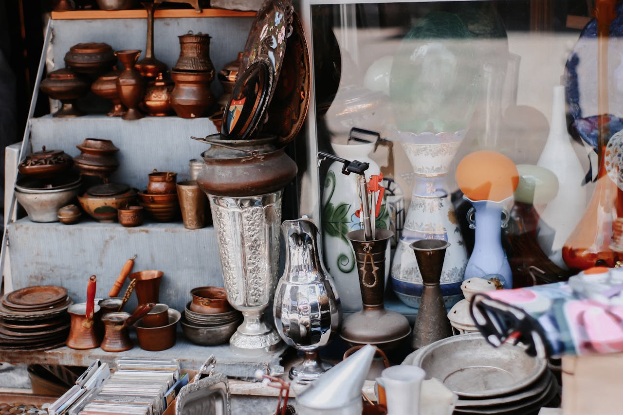 assortment of antique metalware, ceramics, and glass items displayed on shelves and tables at a vintage market