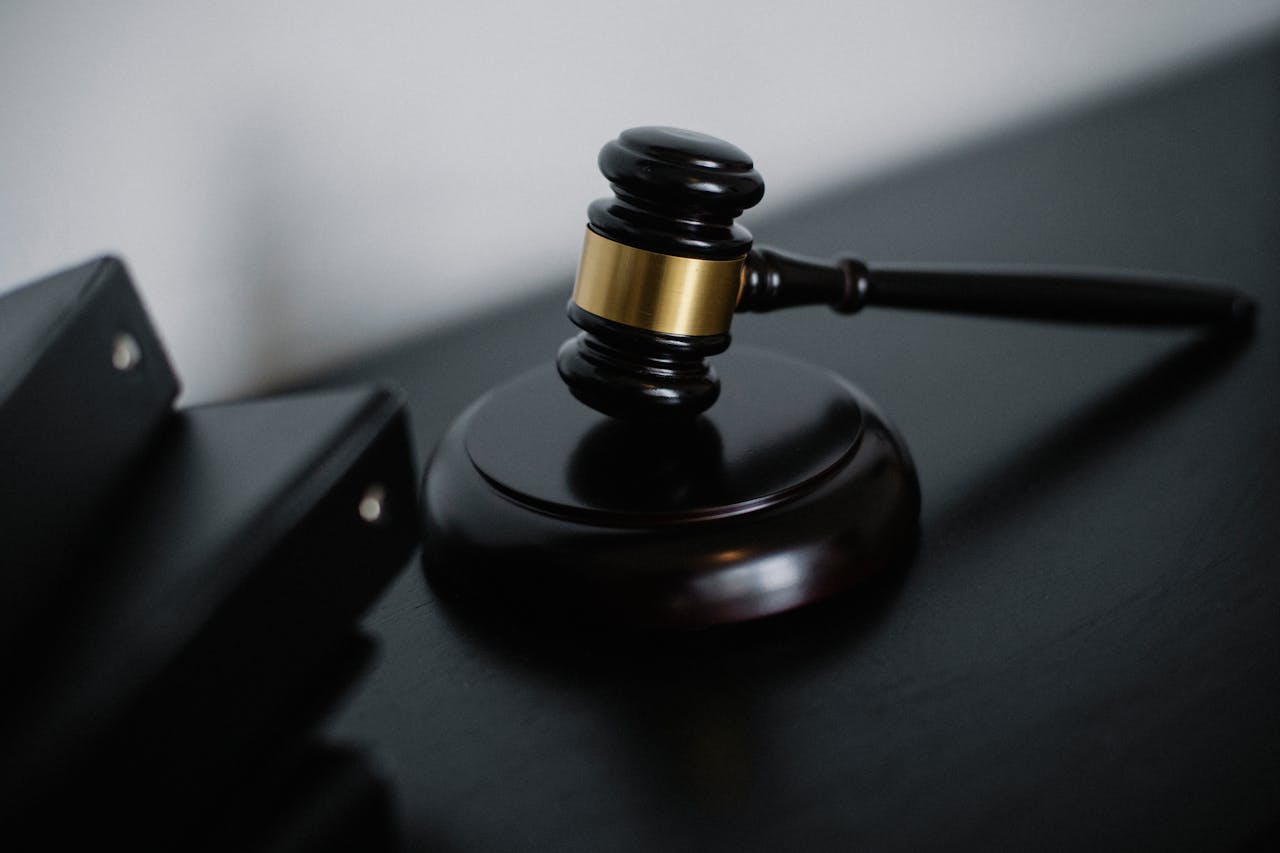 Dark wooden gavel with gold band resting on a round sound block, next to stacked black folders on a dark desk