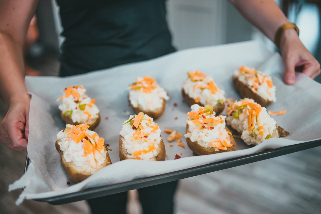 Tray of baked potatoes topped with mashed potato, shredded cheese, and chopped vegetables, held by a person in a kitchen