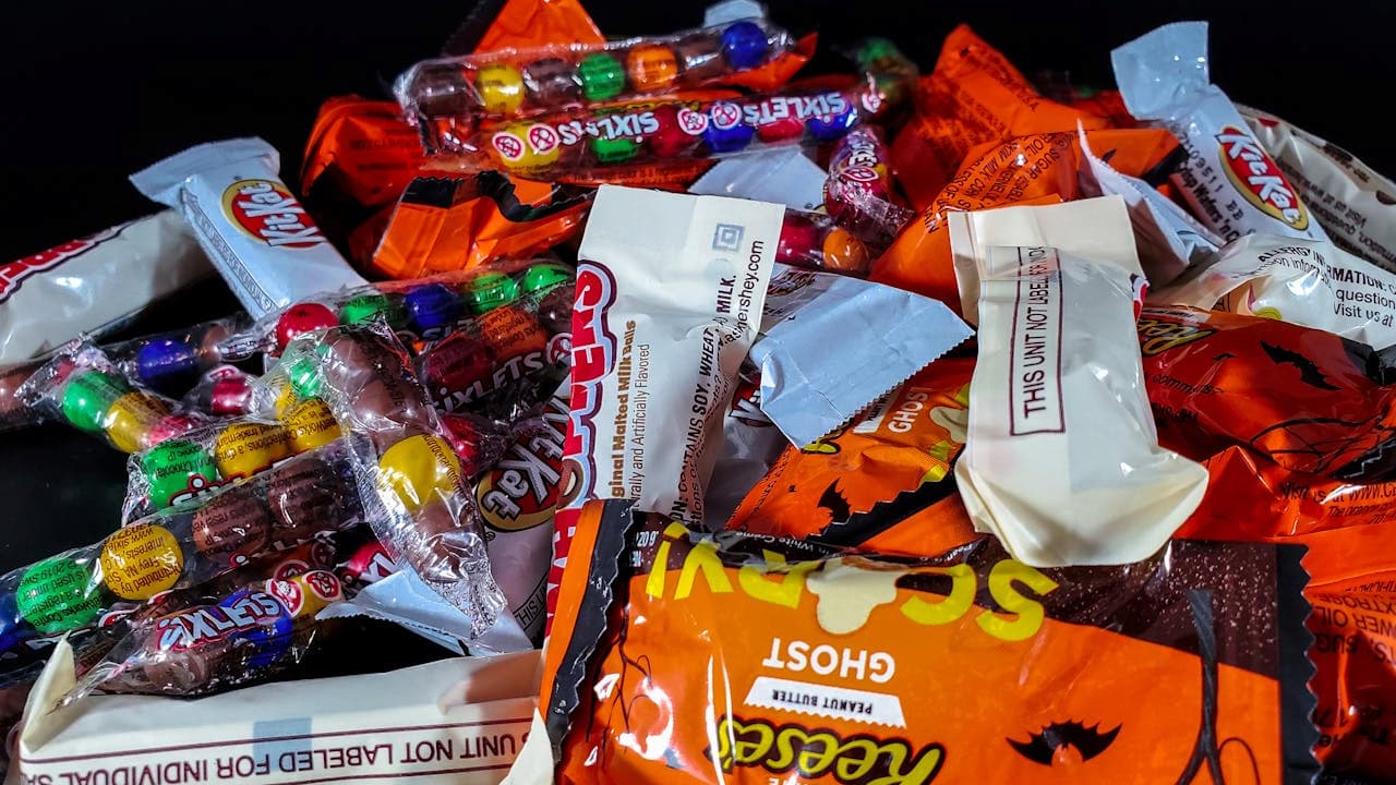 Assorted Halloween candy wrappers, including KitKat, Reese’s, Sixlets, and Snickers, colorful packaging, various sizes, scattered on a dark surface