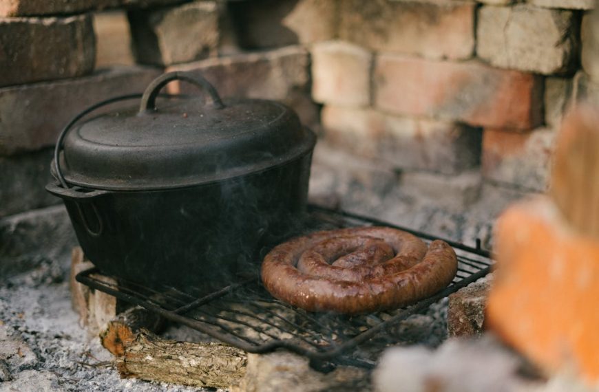 Cast iron Dutch oven and coiled sausage cooking on a rustic outdoor grill, surrounded by brick and firewood, with smoke rising