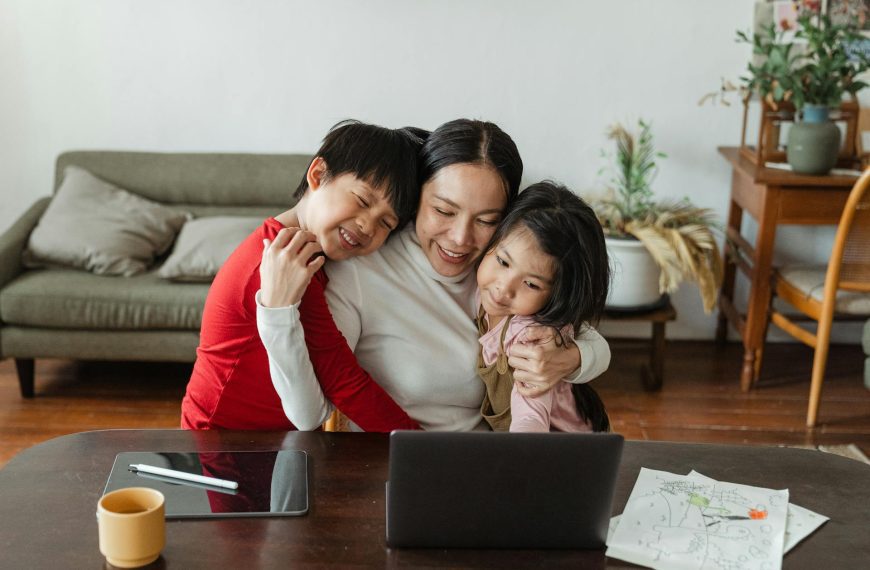 Smiling woman sitting at a table hugging two young children while using a laptop, with a tablet and papers also on the table in a cozy living room