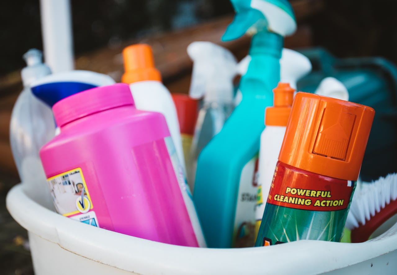 Plastic bin filled with assorted cleaning supplies, spray bottles, detergent containers, colorful packaging, orange and teal accents