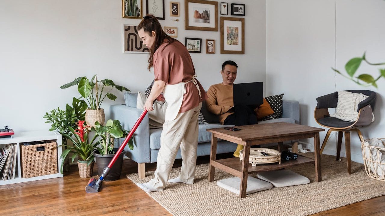 Woman vacuuming wooden floor near couch, man sitting on sofa using laptop, cozy living room with plants and decor
