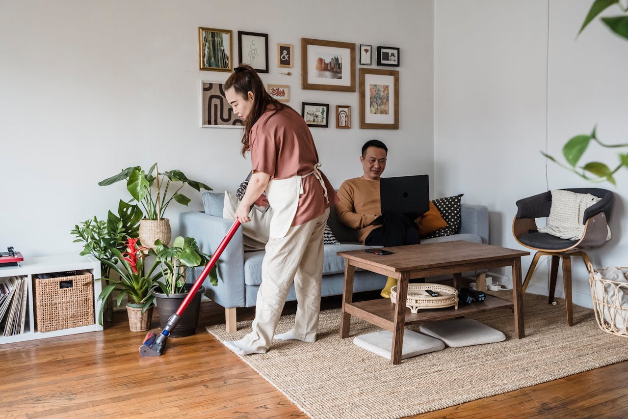 Woman vacuuming wooden floor near couch, man sitting on sofa using laptop, cozy living room with plants and decor