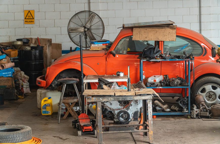 Cluttered garage, disassembled red car, engine parts on workbench, scattered tools, oil containers, industrial fan, warning sign on wall