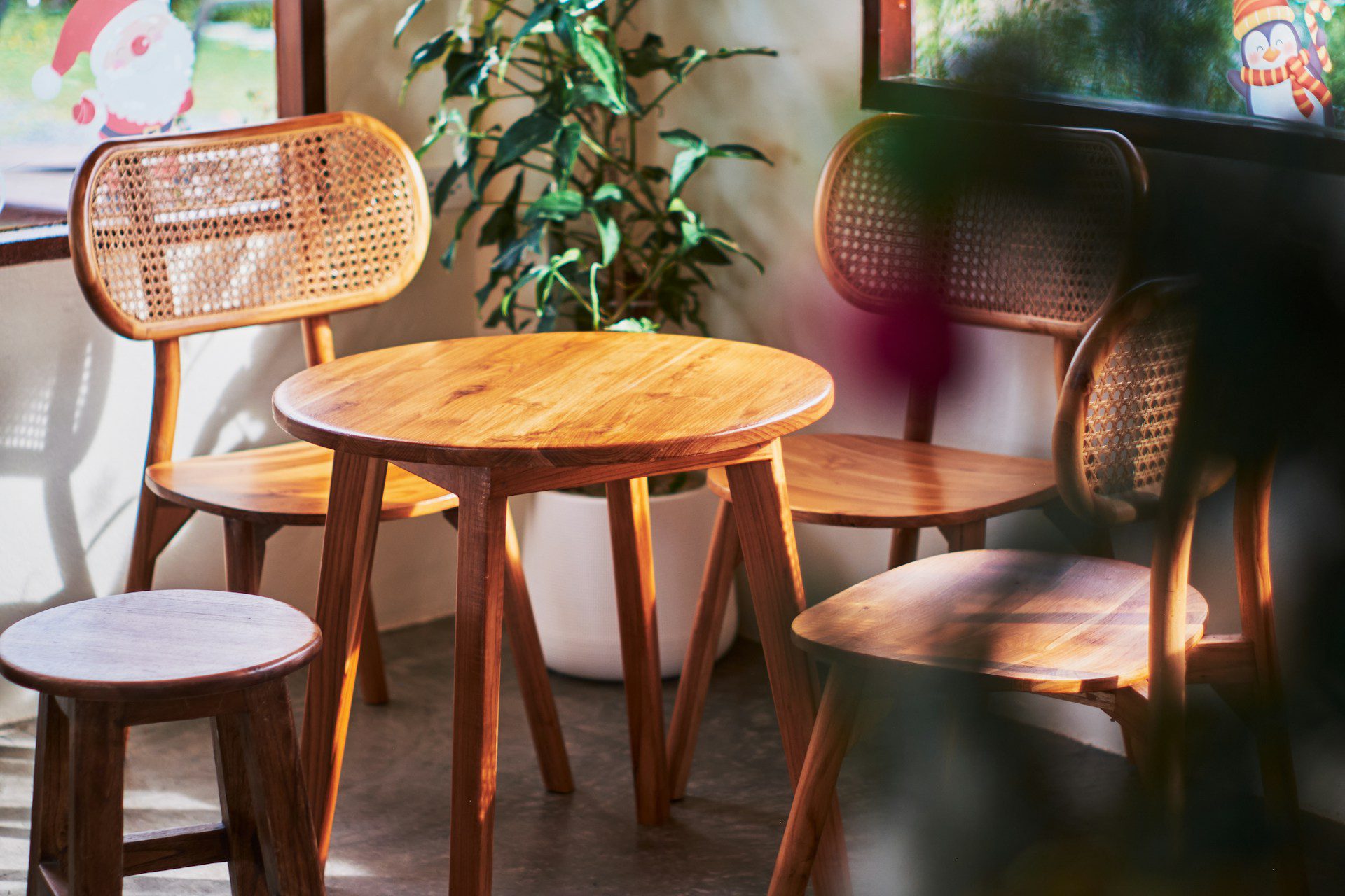 Small round wooden table with three matching wooden chairs, natural sunlight casting warm shadows, indoor plant in the background, cozy and inviting corner, soft focus greenery in the foreground