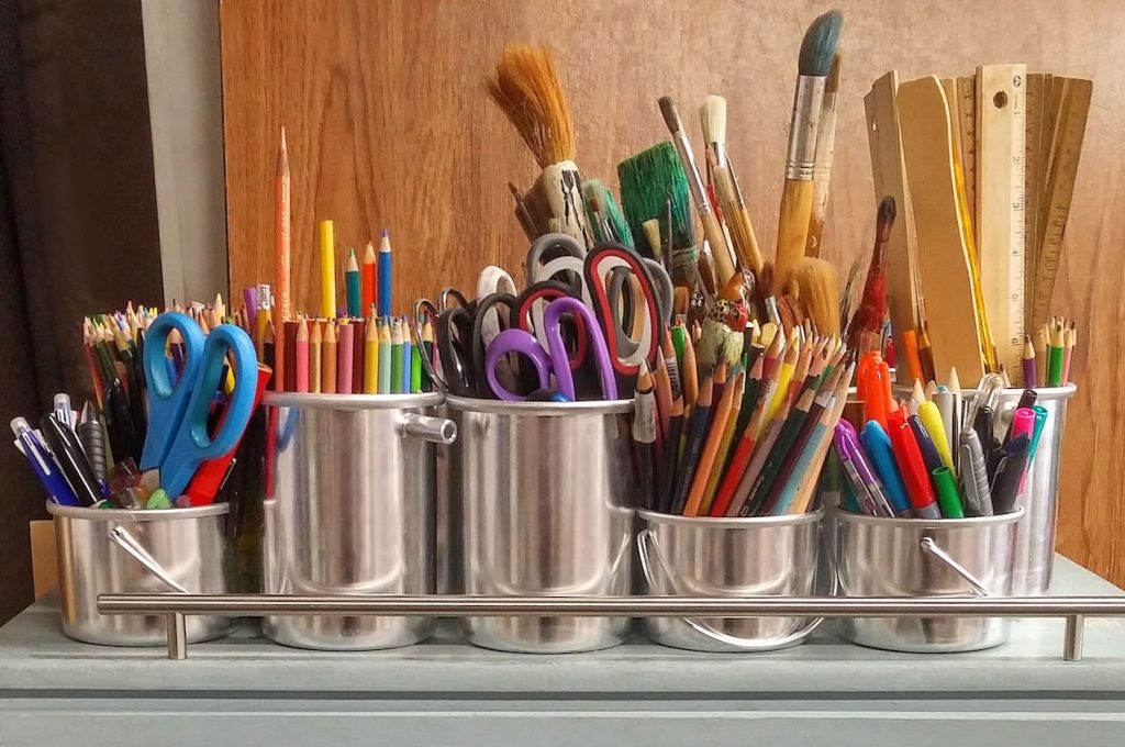 Metal containers filled with colored pencils, scissors, paintbrushes, rulers, pens, markers, organized neatly on a shelf