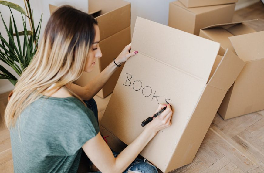 Woman sitting on floor, writing "BOOKS" on cardboard box with black marker, surrounded by boxes, indoor plant nearby