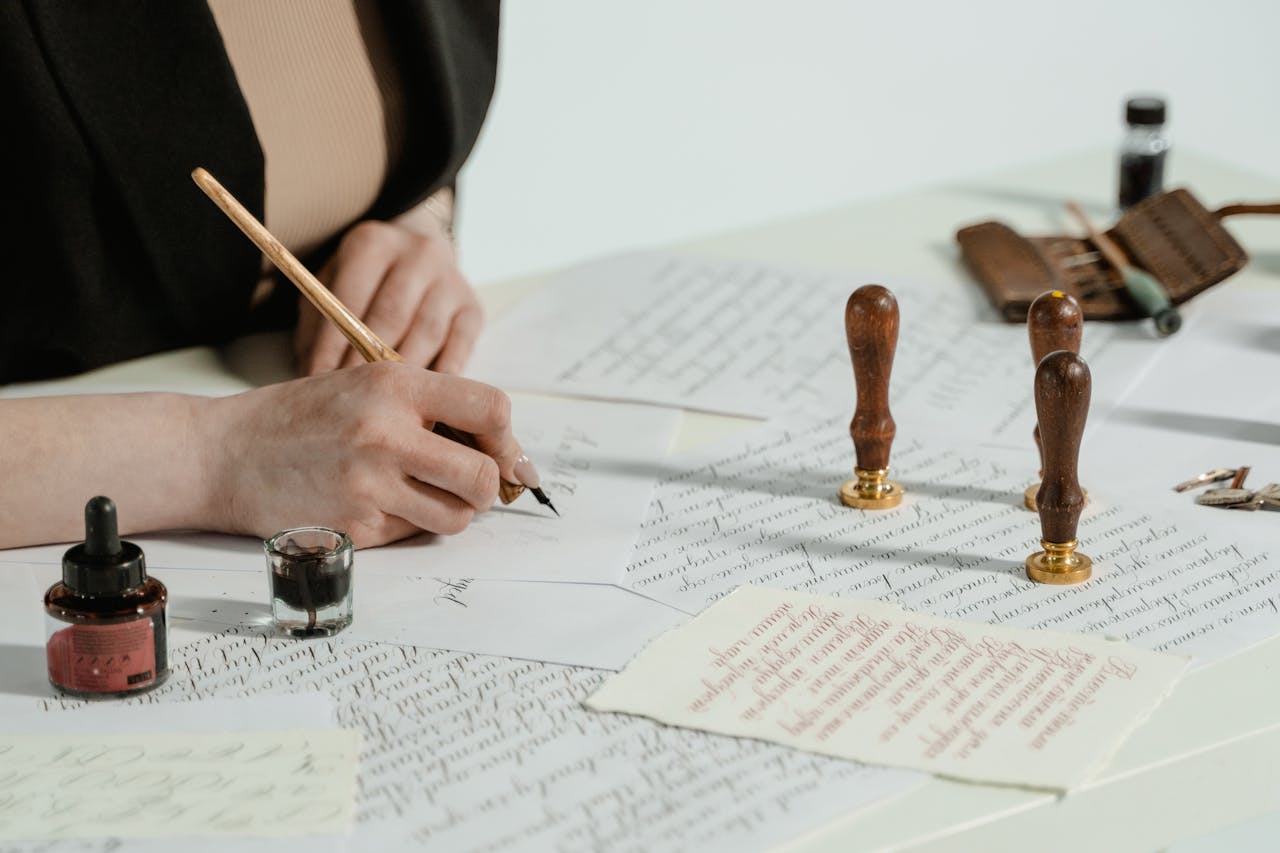 Person using a dip pen to write calligraphy on parchment papers, surrounded by wax seals, ink bottles, and antique writing tools