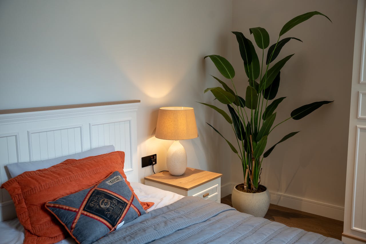 Cozy bedroom corner, white headboard with orange and gray pillows, bedside table with ceramic lamp, large green potted plant, soft lighting, neutral wall and wood flooring