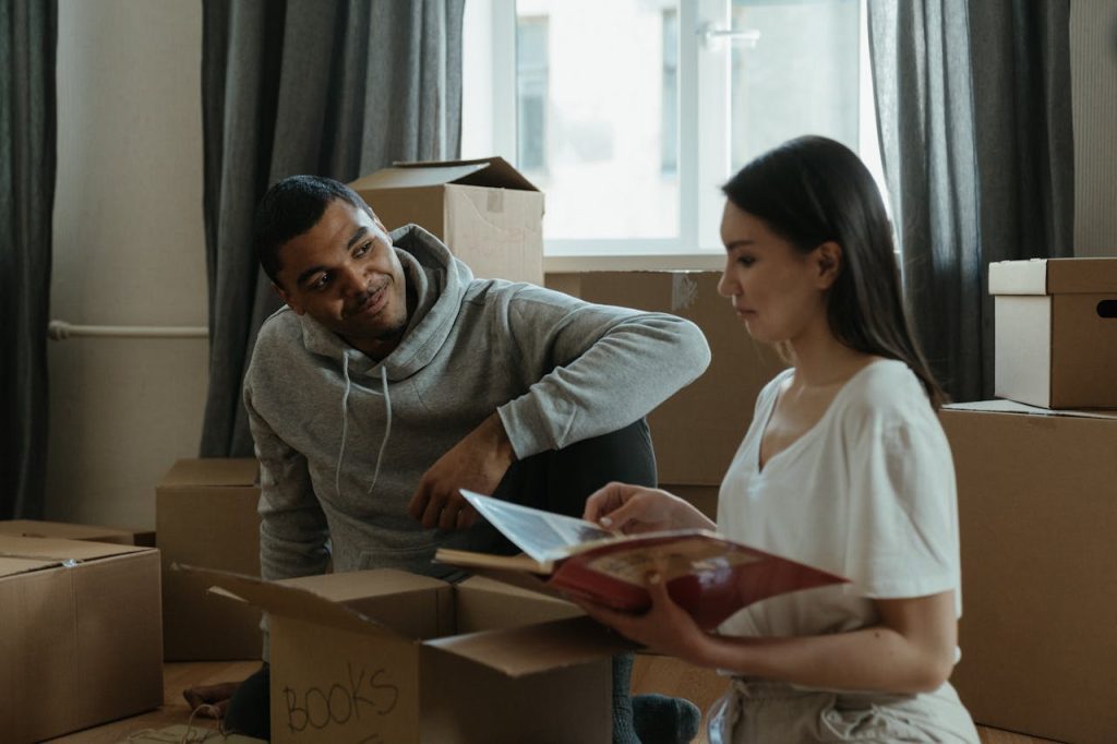 Couple sitting on floor, surrounded by boxes, woman holding open book, man smiling in gray hoodie, daylight through window