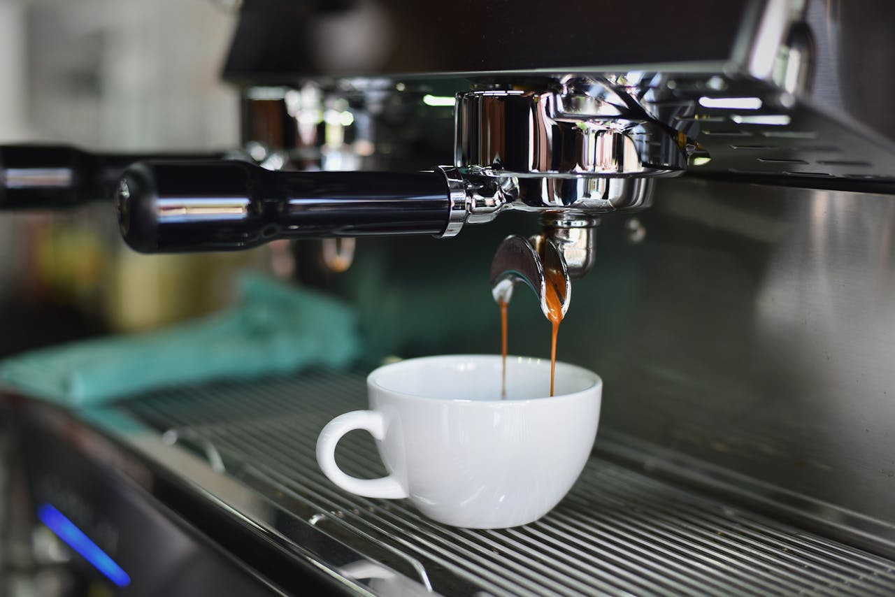 Close-up of espresso machine brewing coffee into a white ceramic cup. Two streams of dark espresso pour from a portafilter with black handle, showing crema formation over metal drip tray in shallow focus