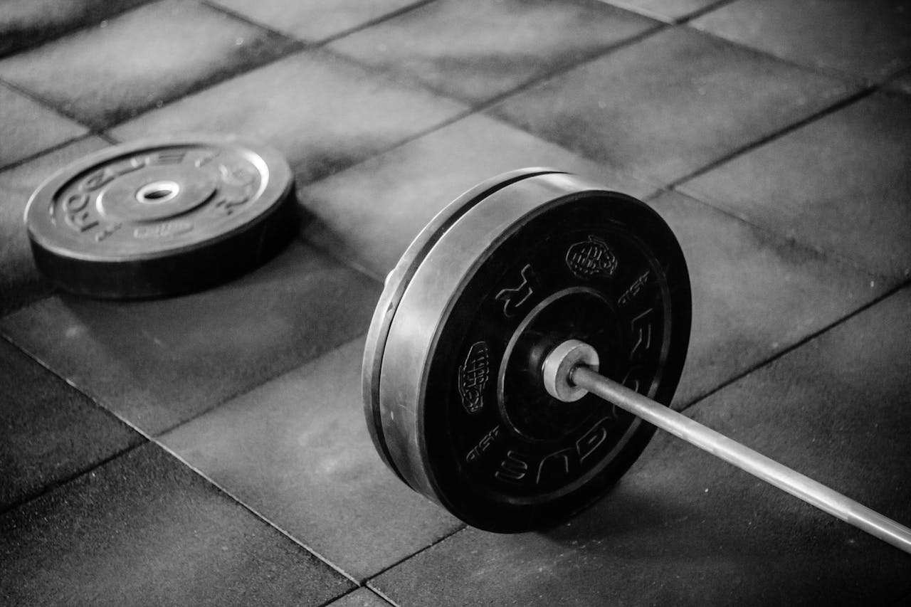 Black barbell with weight plates on a rubber gym floor, extra plate lying nearby, monochrome lighting