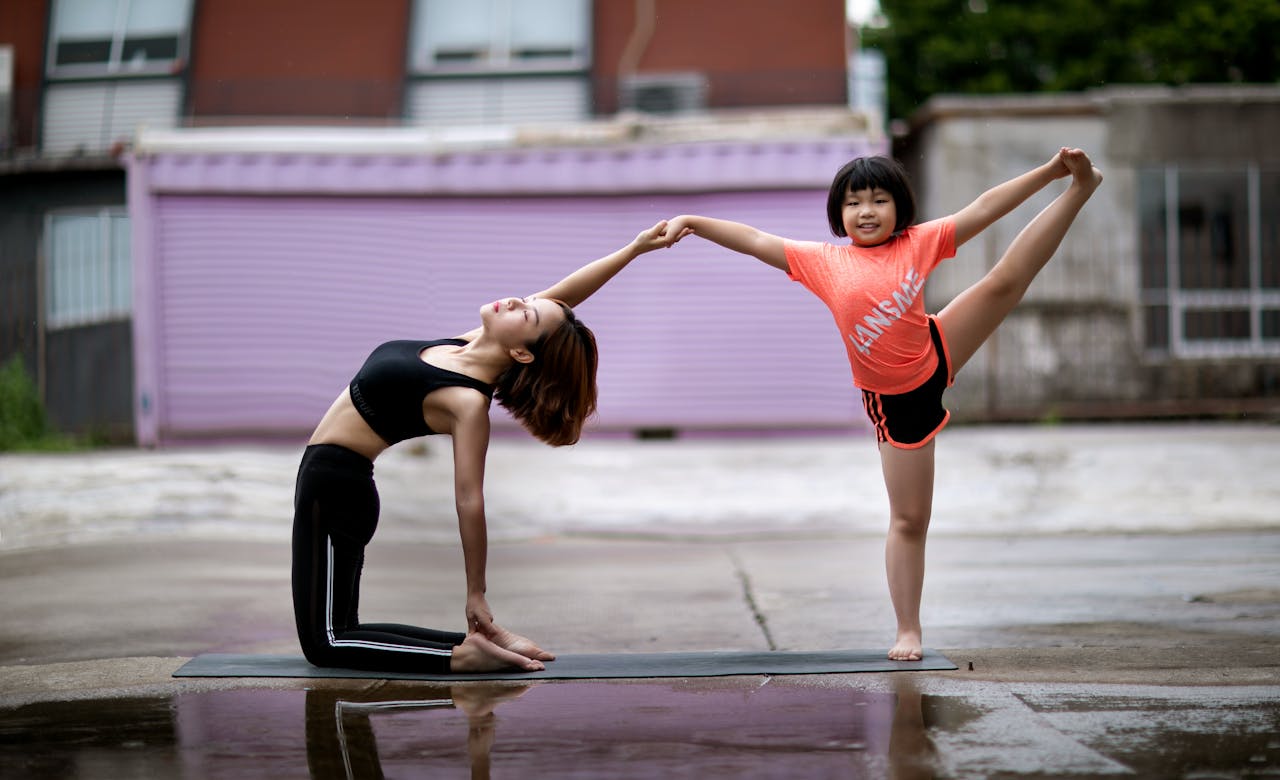 Woman and girl doing yoga poses while holding hands on a wet concrete surface
