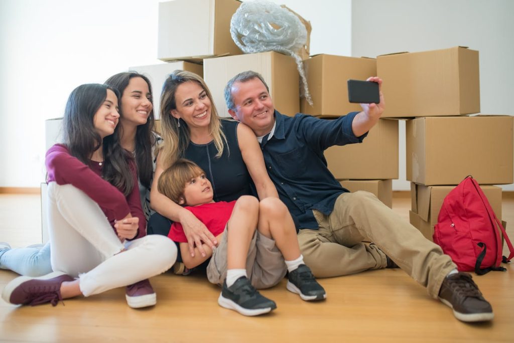 Smiling family of five, sitting on floor, taking selfie, surrounded by cardboard boxes, moving day, happy expressions, casual clothes