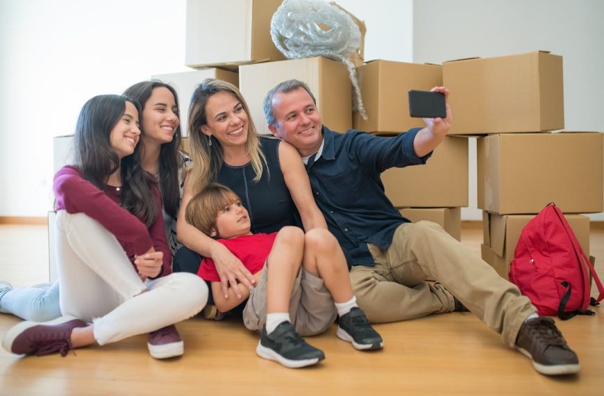 Smiling family of five, sitting on floor, taking selfie, surrounded by cardboard boxes, moving day, happy expressions, casual clothes
