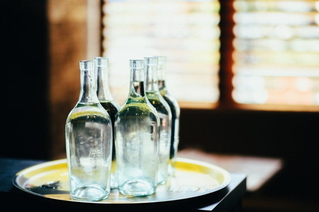 six clear glass water bottles, placed on round metal tray, backlit by sunlight through window blinds, indoor setting